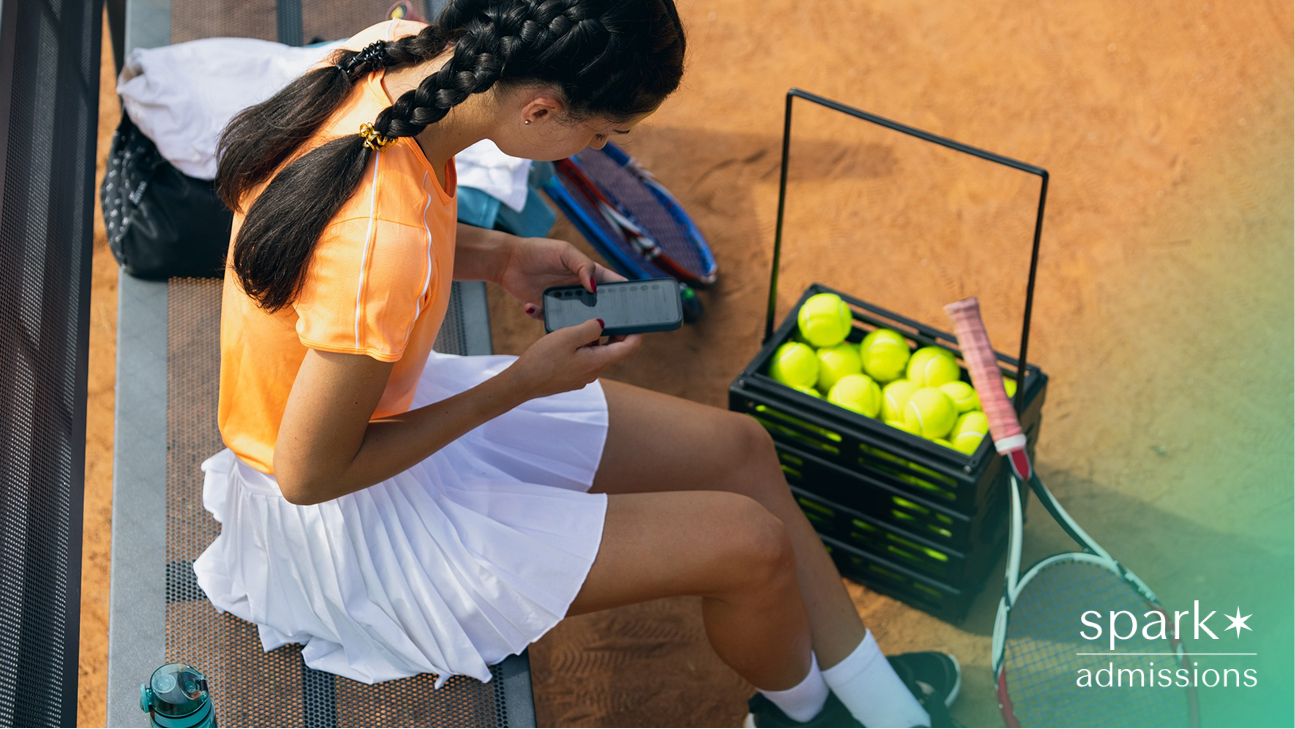 Teen tennis player sitting on a bench checking her phone next to a basket of tennis balls