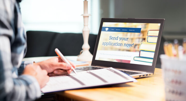 A man filling out a paper application, while sitting in front of a laptop that states "Send your application now!".