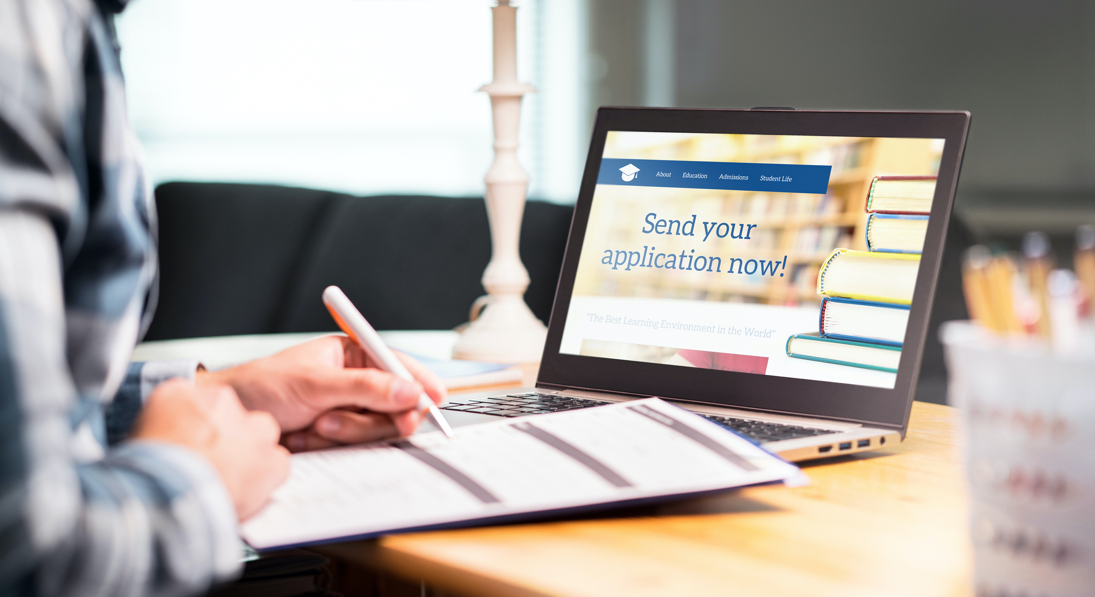 A man filling out a paper application, while sitting in front of a laptop that states "Send your application now!".