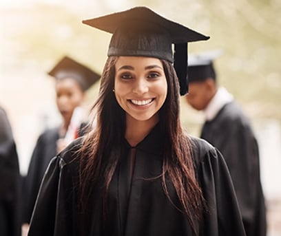 A high school girl graduate in a cap and gown with classmates in background