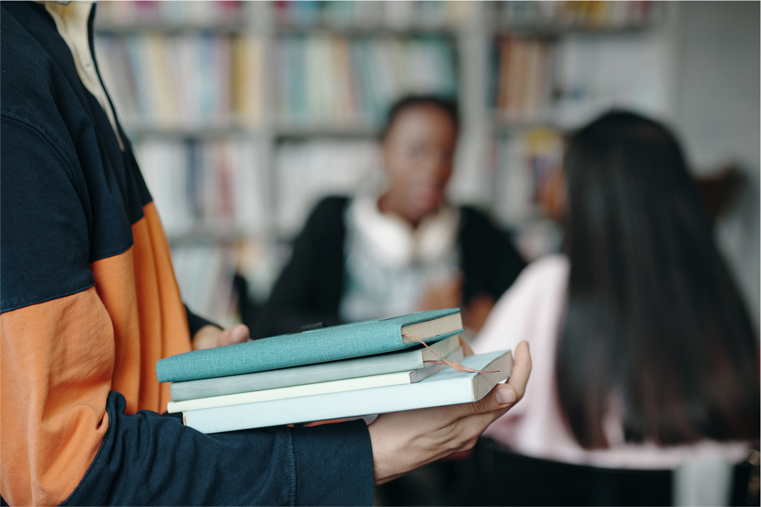 Close-up of a person holding books with students talking in the background at a library