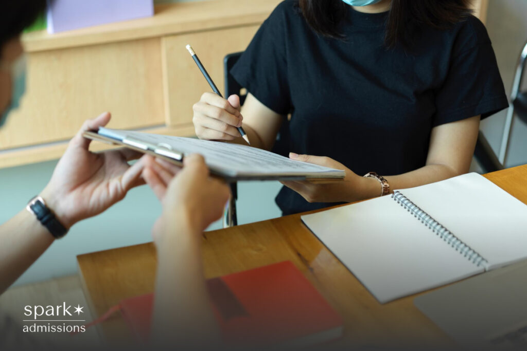 Two students in masks sit at a table reviewing a clipboard together, with notebooks nearby
