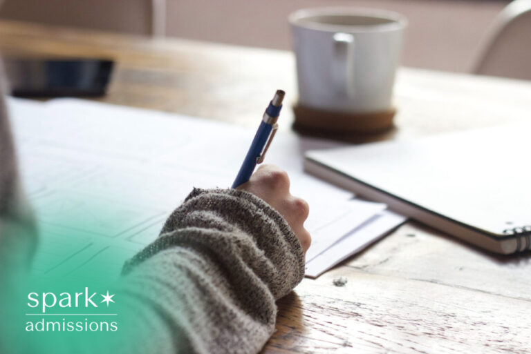 Student writing notes at a wooden desk with a notebook, planner, and coffee mug nearby.
