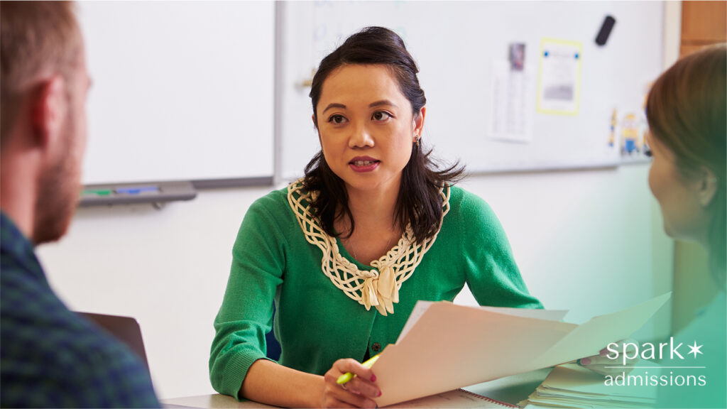 Admissions counselor in a green sweater talking to two people across a desk, holding paperwork