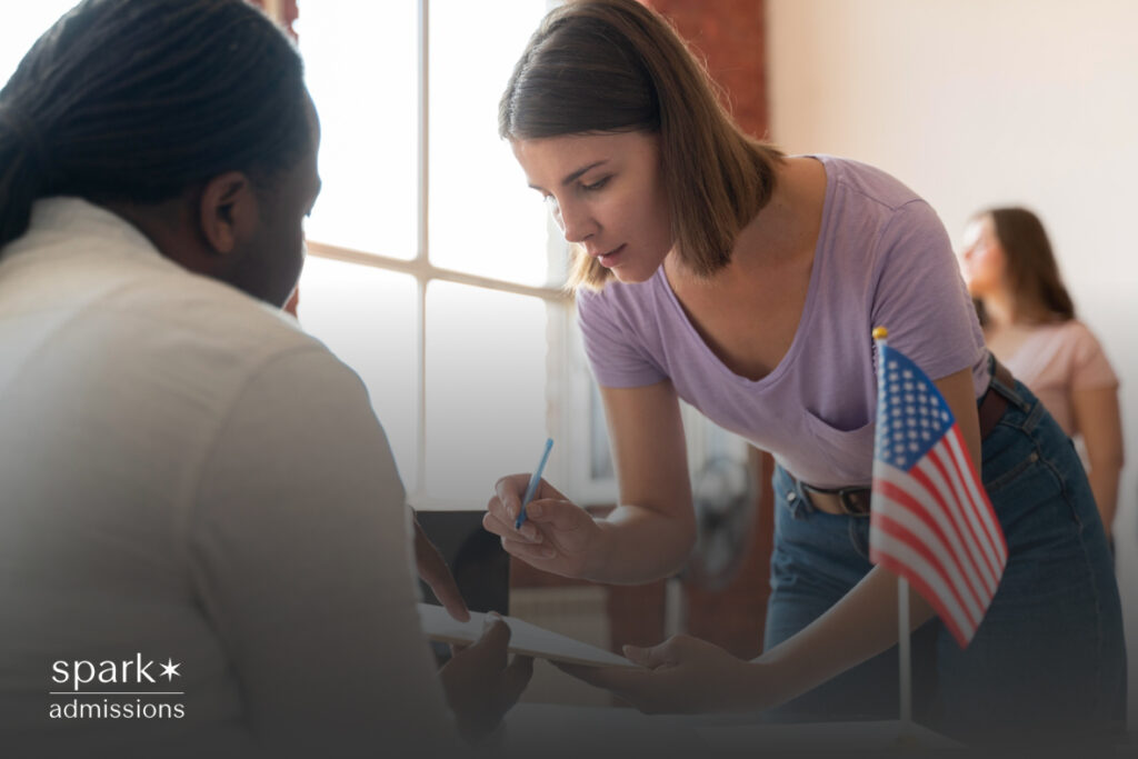 A young woman leans over to help someone fill out a form next to a small American flag on a desk