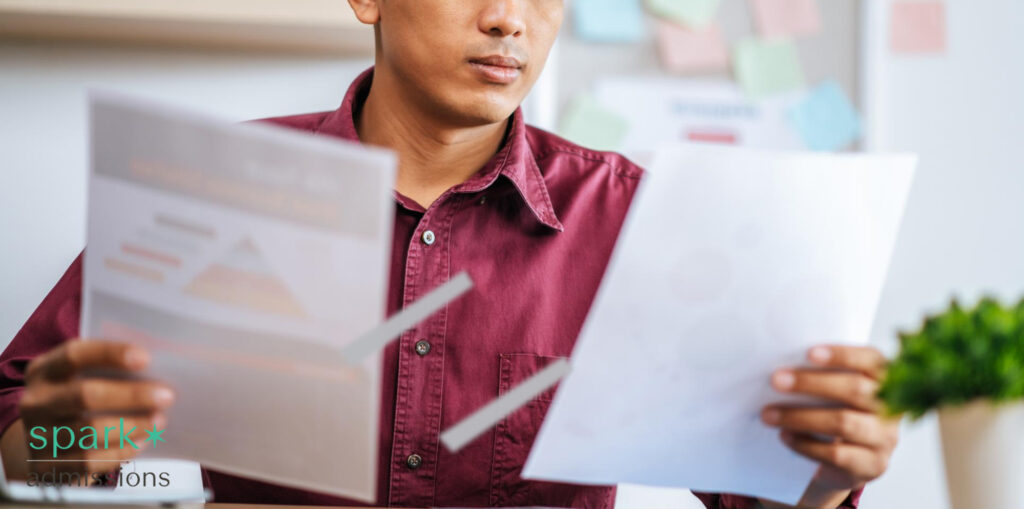 Man in a maroon shirt comparing two printed documents at a desk, with Spark Admissions logo in the corner