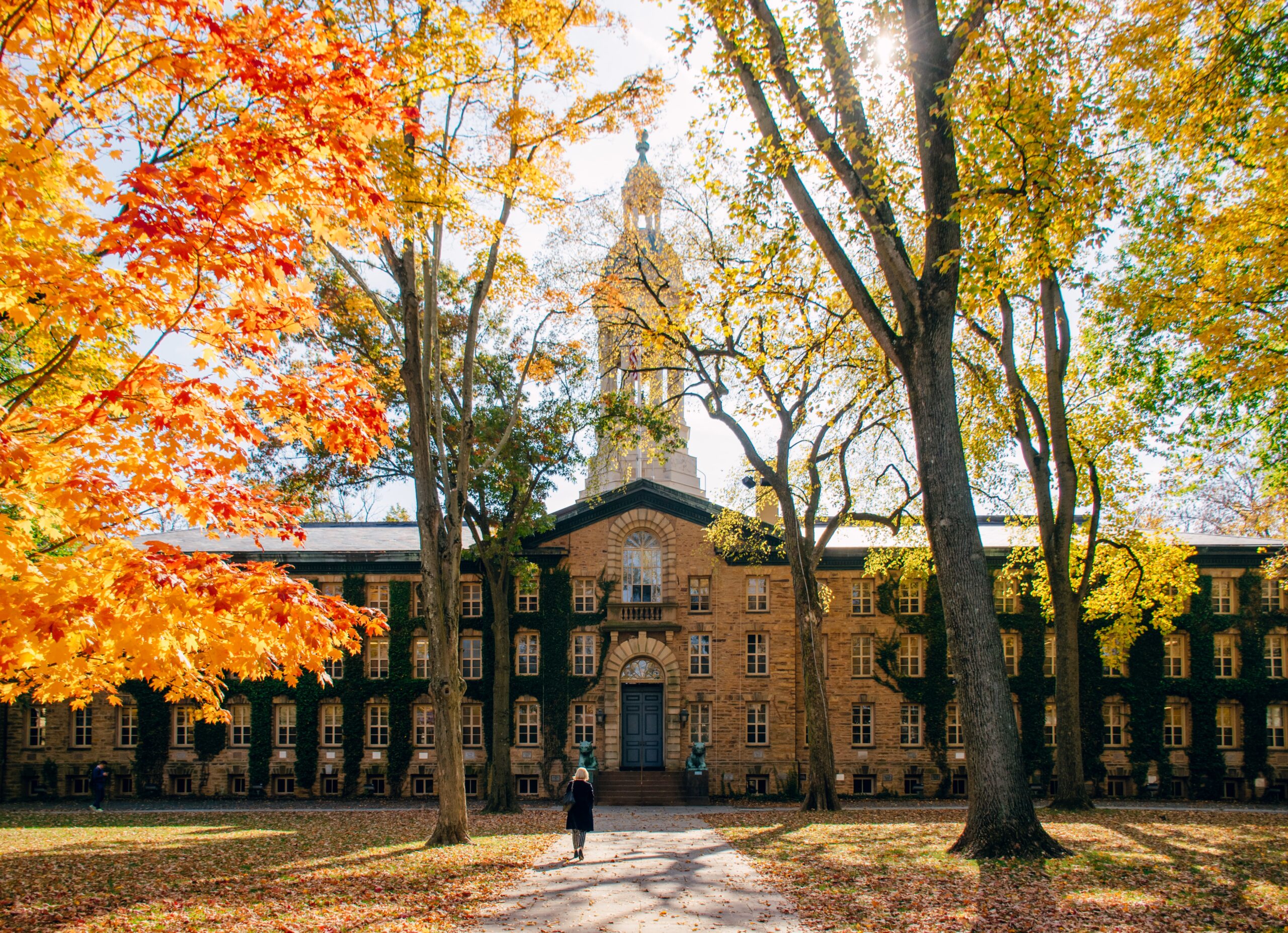 A student walks toward a historic university building surrounded by colorful fall trees