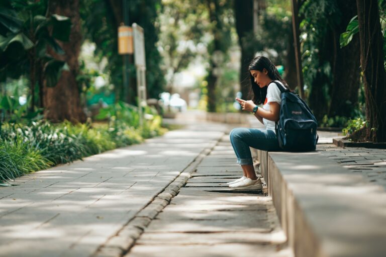 A student with a backpack sitting on a low ledge in a tree-lined walkway, looking at her phone
