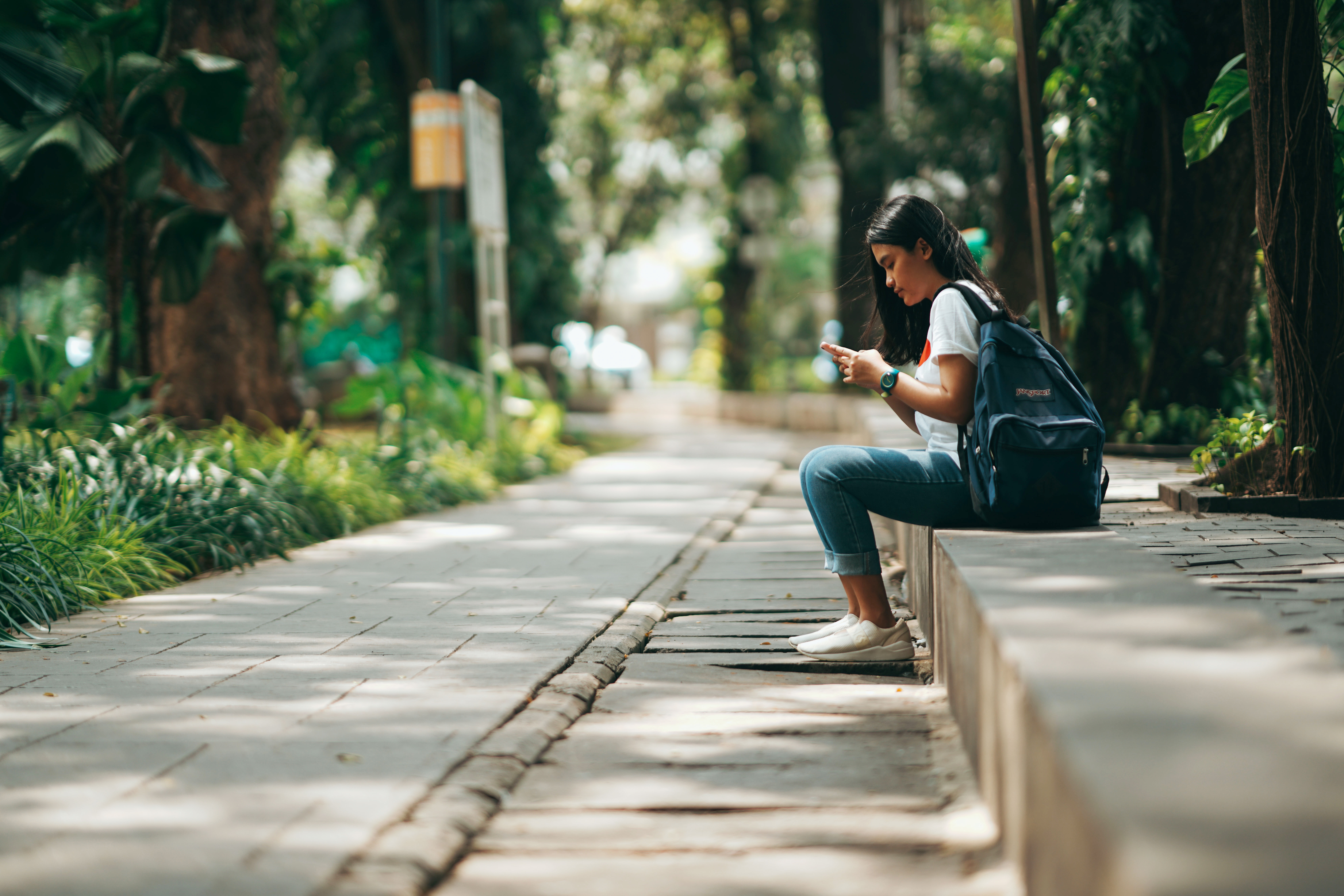 A student with a backpack sitting on a low ledge in a tree-lined walkway, looking at her phone