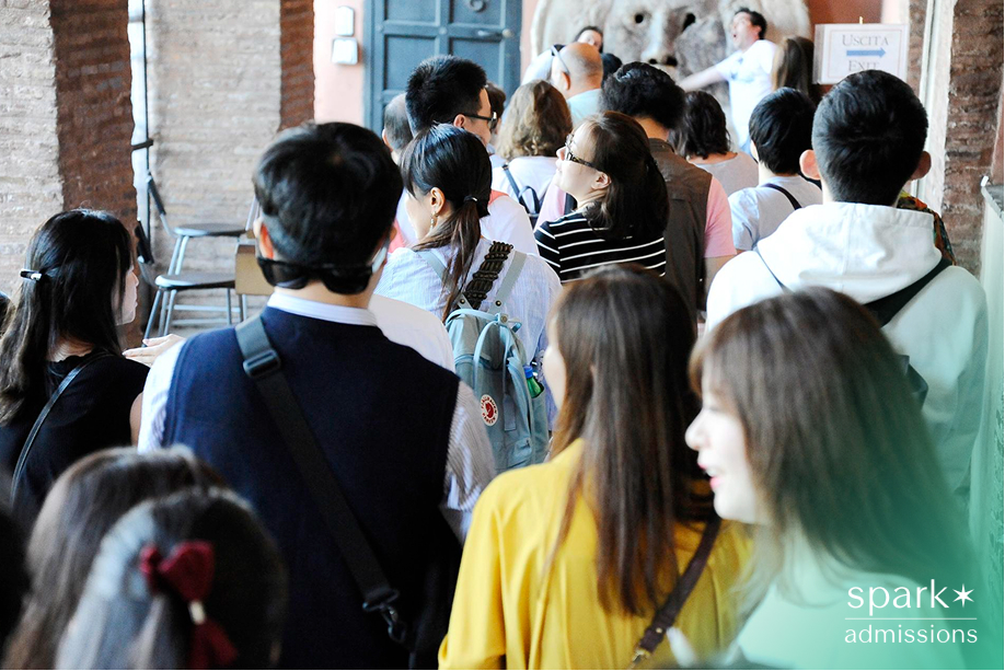 Large group of prospective students standing in line at a college admissions or campus event, viewed from behind in a historic building.