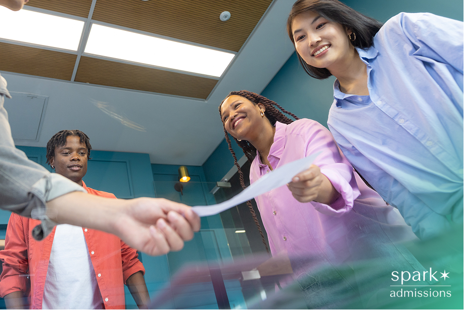 Three diverse students smiling as one hands over a paper in a modern office setting, representing college admissions success or acceptance.