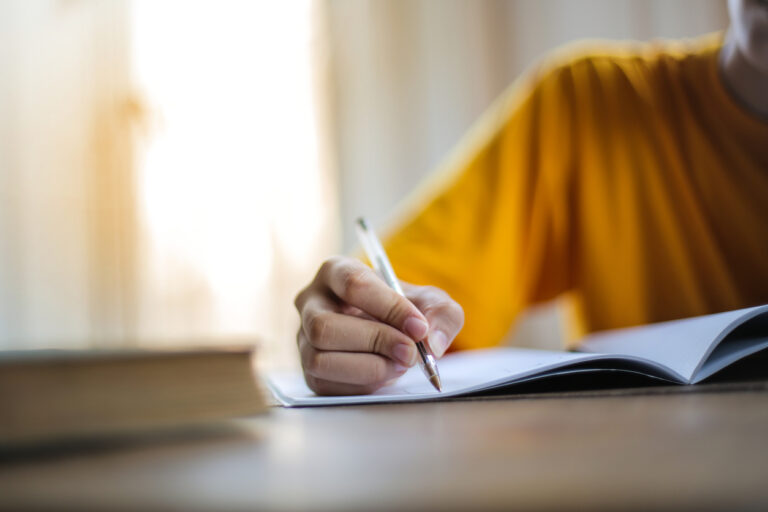 A student in a yellow shirt writing in a notebook at a wooden desk near a window