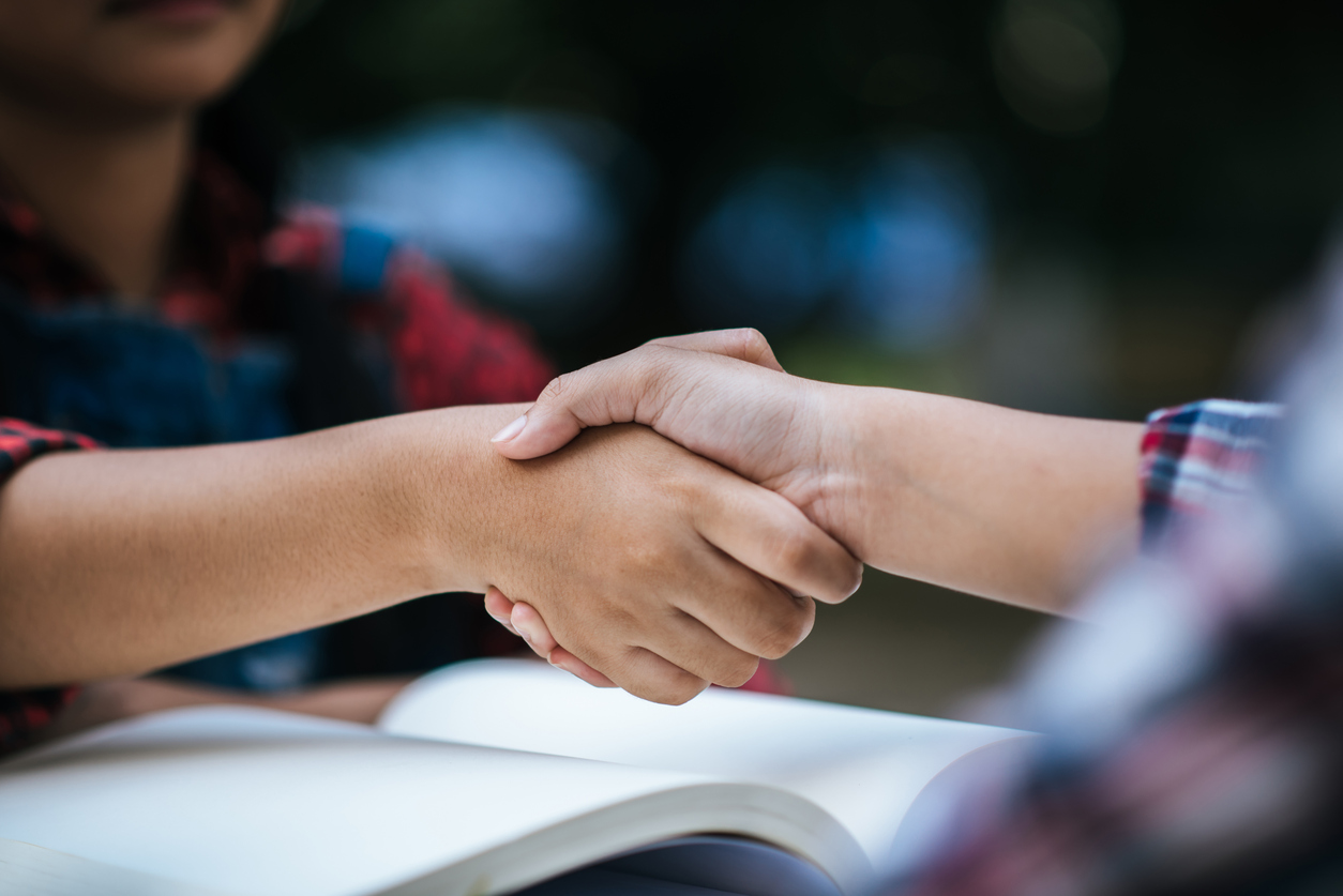 Two students shaking hands over an open book during an outdoor study session