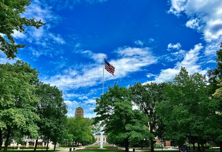An American flag waves over a tree-lined University of Michigan, The Ross School of Business campus lawn on a sunny day, creating a classic collegiate atmosphere