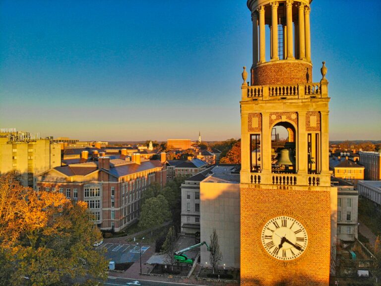 UNC Chapel Hill’s bell tower stands tall above the campus at golden hour, with warm light casting shadows across the historic brick buildings and autumn foliage