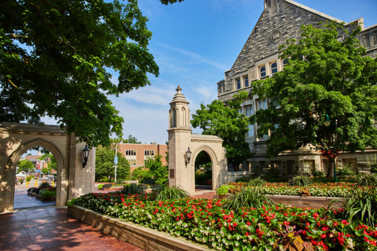 Indiana University’s iconic Sample Gates arch is surrounded by vibrant red flowers and lush greenery on a sunny day, welcoming visitors to a traditional college campus