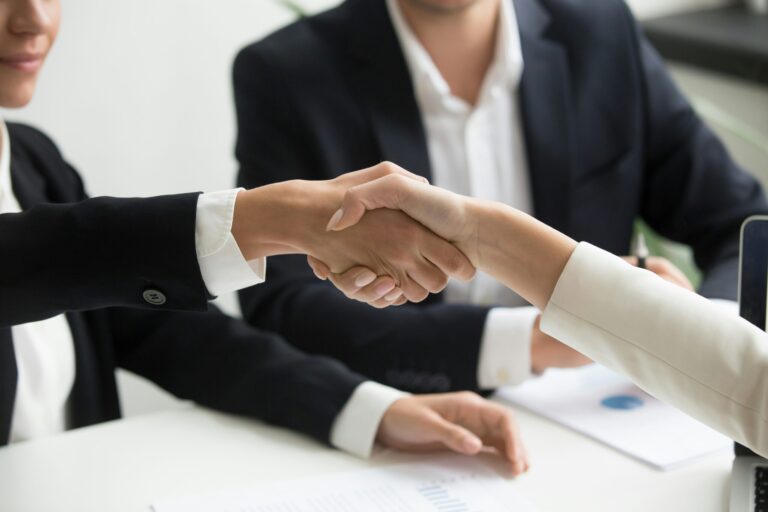 Two professionals shake hands across a desk in a corporate setting, symbolizing agreement, collaboration