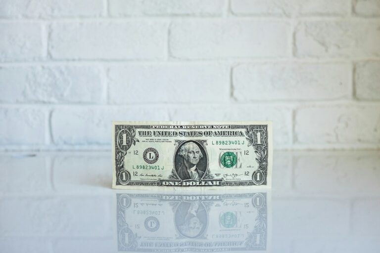 A single U.S. dollar bill standing upright against a white brick wall with a reflective surface