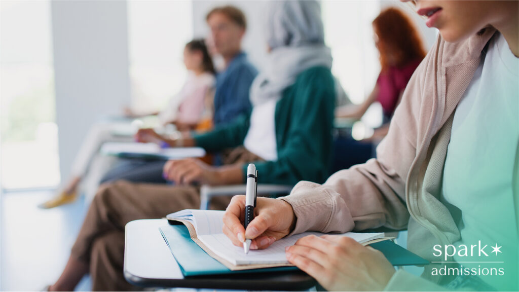 A student writing in a notebook during class, with other students blurred in the background