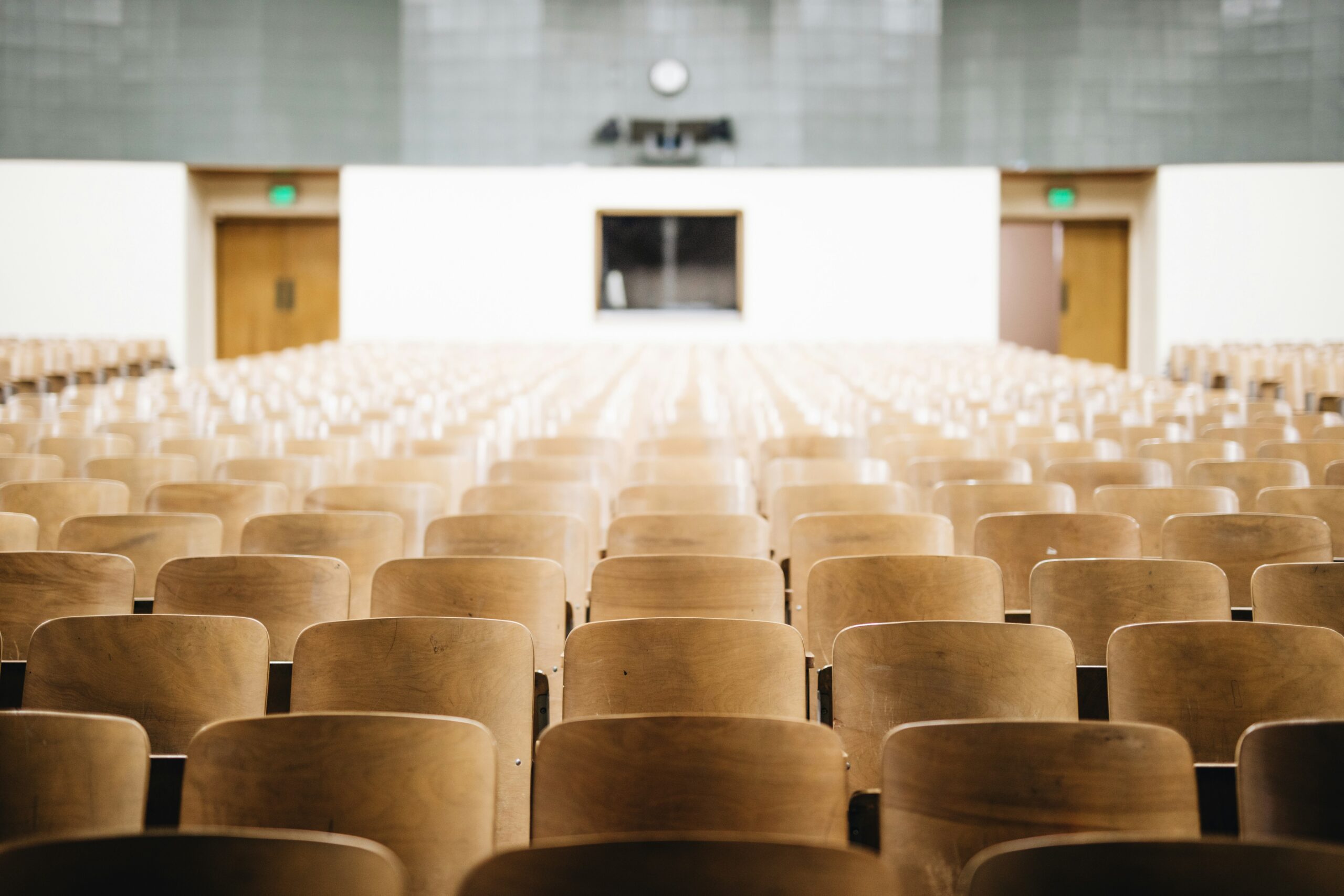Empty wooden chairs in a large lecture hall facing a podium and whiteboard
