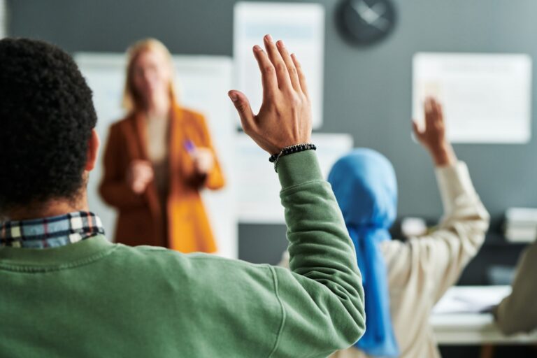 Two students raise their hands to participate while a teacher leads a discussion in front of a classroom