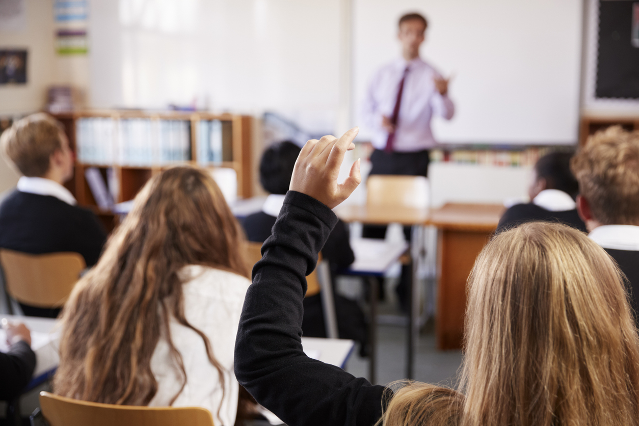 Female student raising their hand in a classroom while a teacher stands at the front
