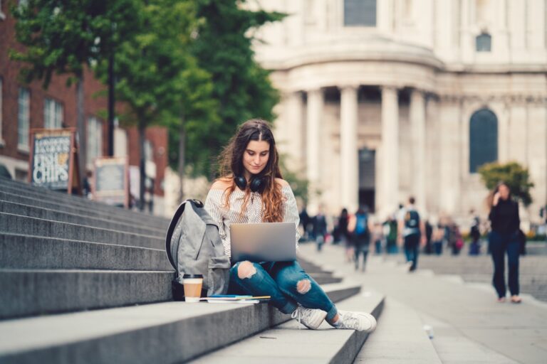 A college student types on a laptop while sitting on stone steps with coffee and a backpack nearby