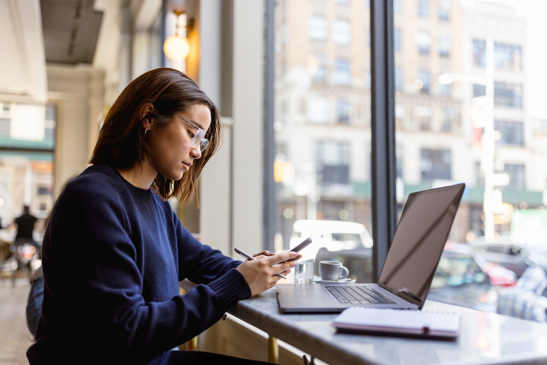 A student in glasses sits by a window in a cafe, working on her phone with a laptop open in front of her