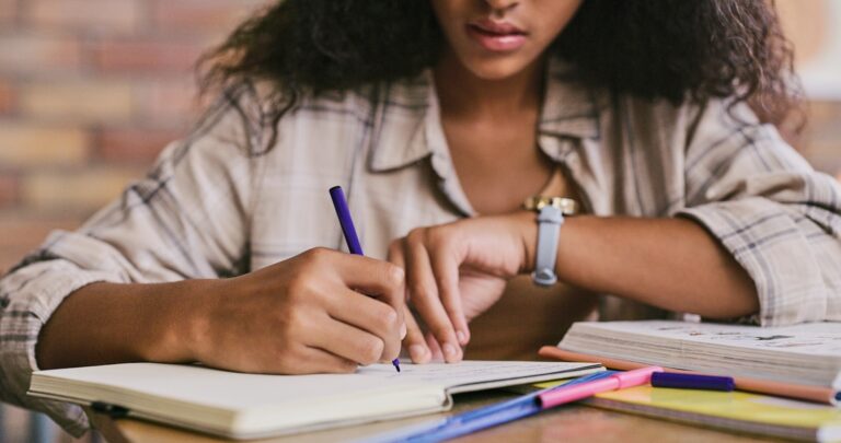 A student sits at a desk, writing in a notebook with colorful pens and study materials spread around