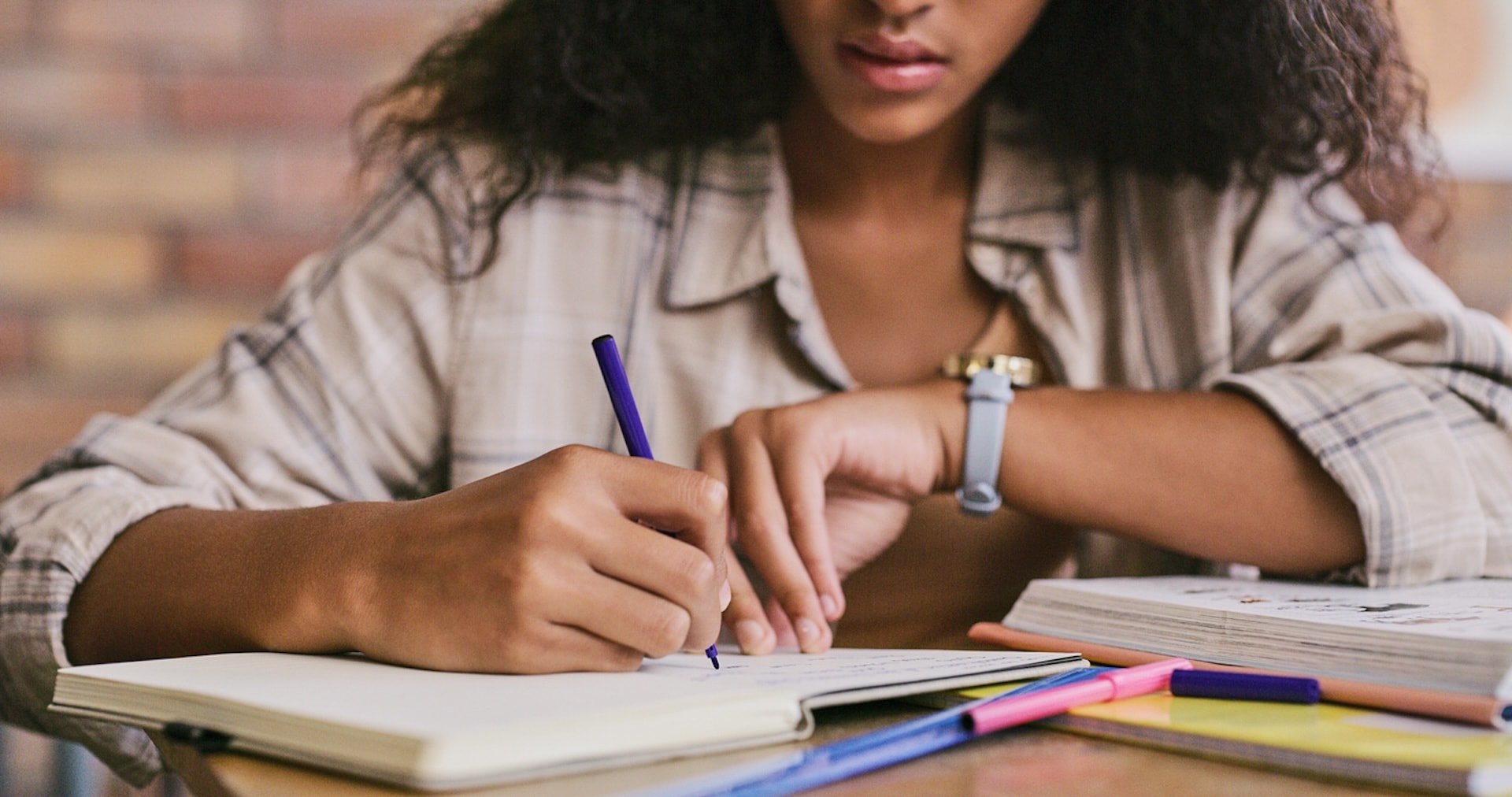 A student sits at a desk, writing in a notebook with colorful pens and study materials spread around