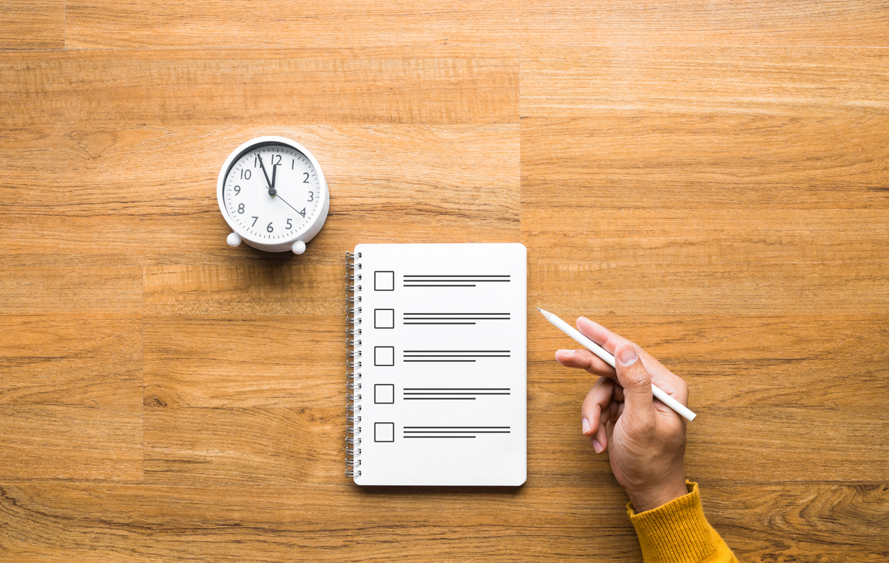 Hand holding a pen above a checklist notebook with a clock on a wooden desk