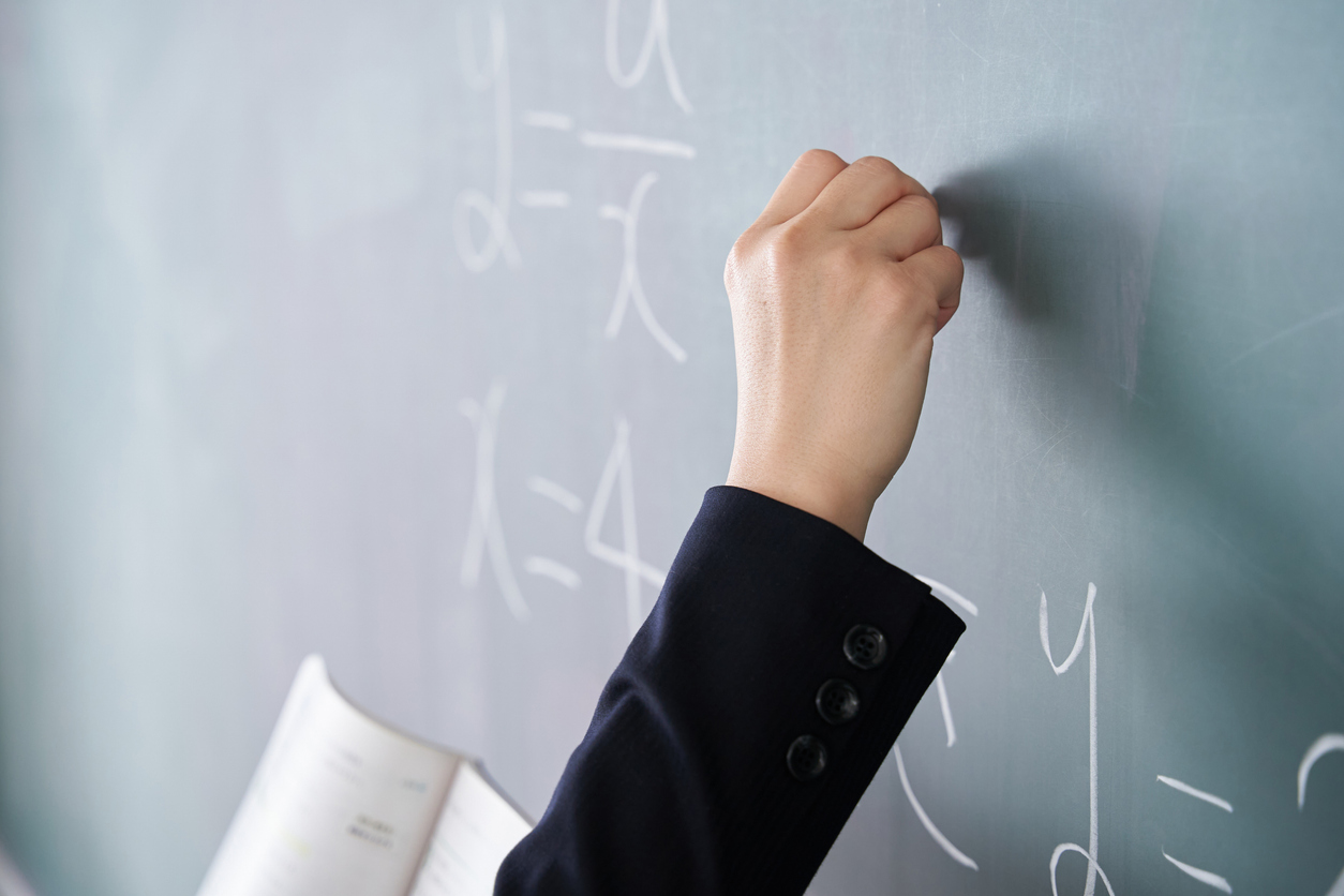 A teacher's hand writing algebraic equations on a green chalkboard while holding an open textbook