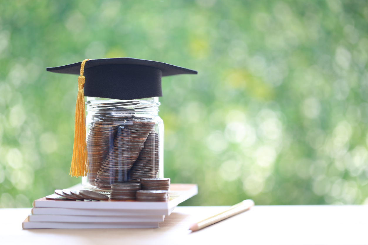 A graduation cap placed on a jar full of coins stacked on top of books, symbolizing saving for education