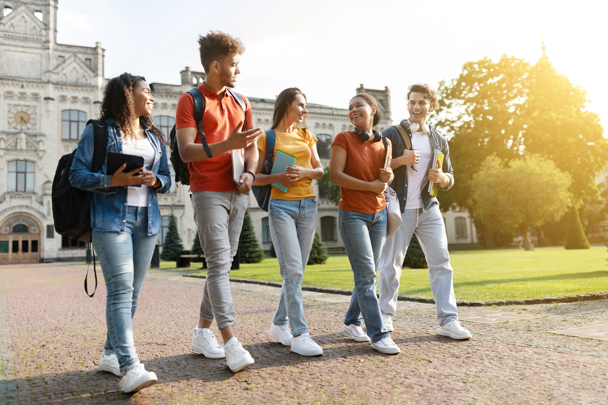 Five students walk together outside a college building, smiling and carrying books