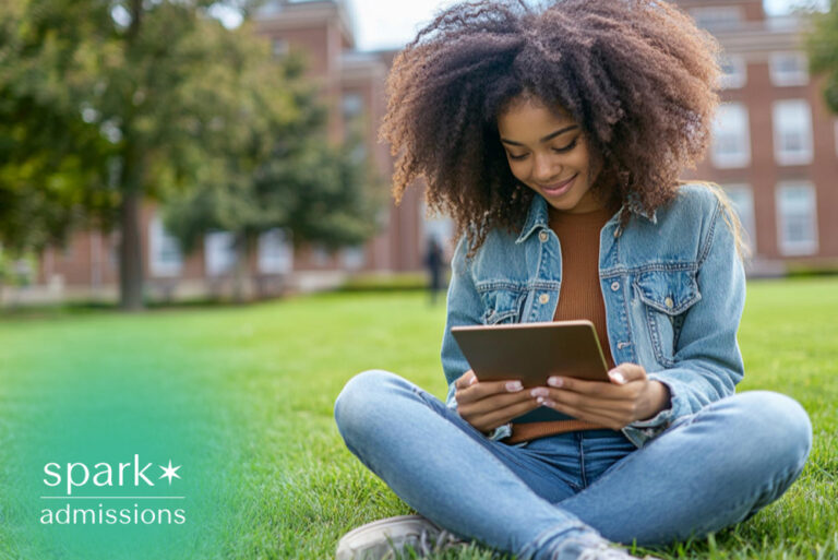 Smiling college student sitting on a campus lawn using a tablet, studying outdoors in front of a university building on a sunny day.