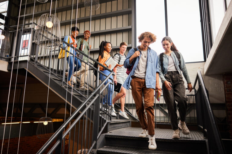 A group of smiling students walk down a stairwell inside a modern school building, holding books and backpacks