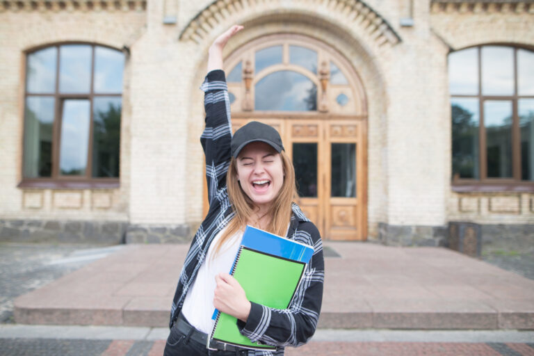 A student raises her arm in celebration while standing outside a university building