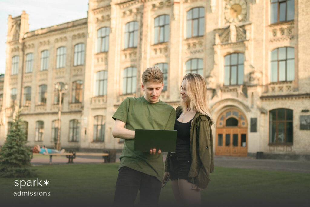 Two students stand in front of a historic university building, smiling and looking at a laptop