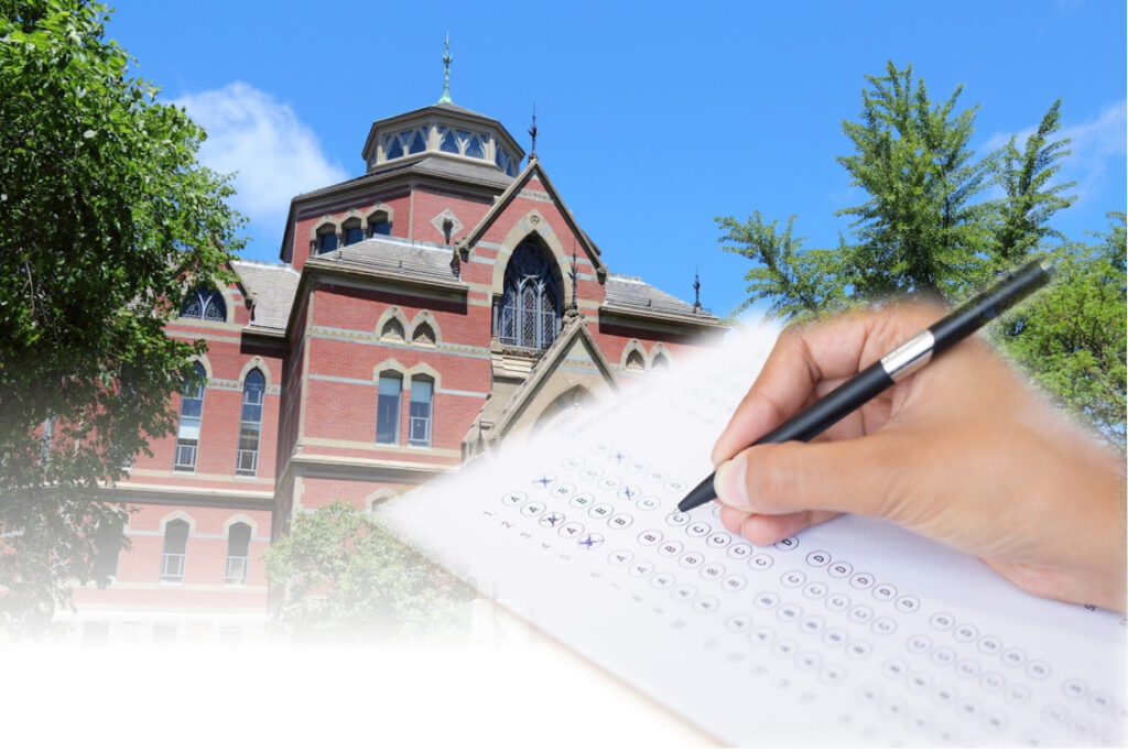 A hand fills out a bubble answer sheet, overlaid in front of a red-brick college building and blue sky