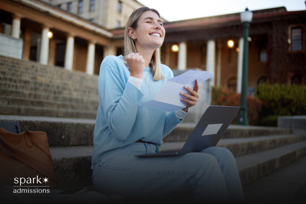 A woman celebrates after reading a college acceptance letter while seated with her laptop on campus stairs