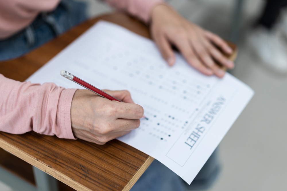 A student sits at a desk filling out a multiple-choice answer sheet with a pencil
