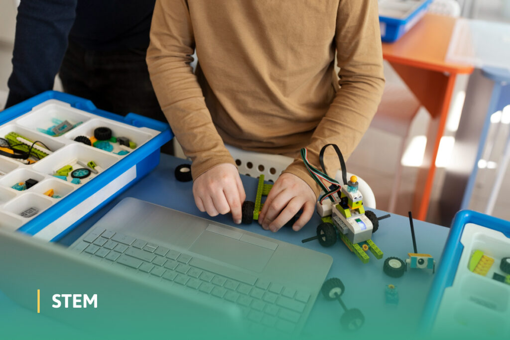 A child builds a small wheeled robot using STEM tools and a laptop on a classroom table