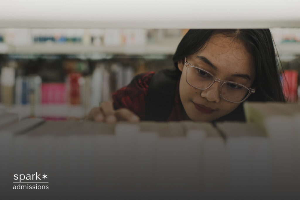 A student wearing glasses reaches for a book on a crowded library shelf