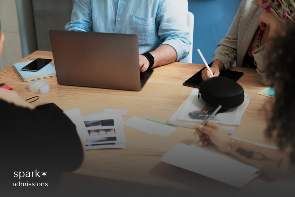 Four individuals sit at a wooden table with a laptop, notes, and printed materials spread out