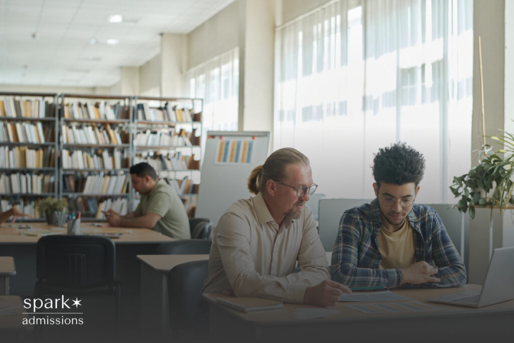 An older man and a younger student go over papers together in a library filled with bookshelves and natural light