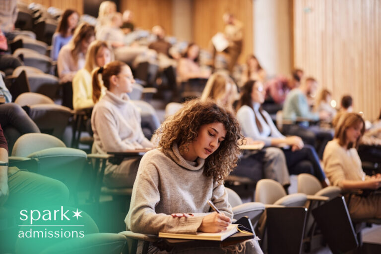 College student taking notes during a university lecture in a large auditorium classroom with other students listening and participating in class.