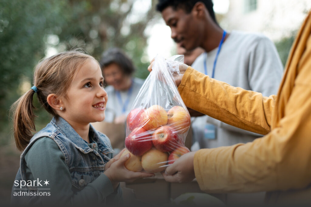 A young girl smiles as someone hands her a plastic bag filled with apples at an outdoor event