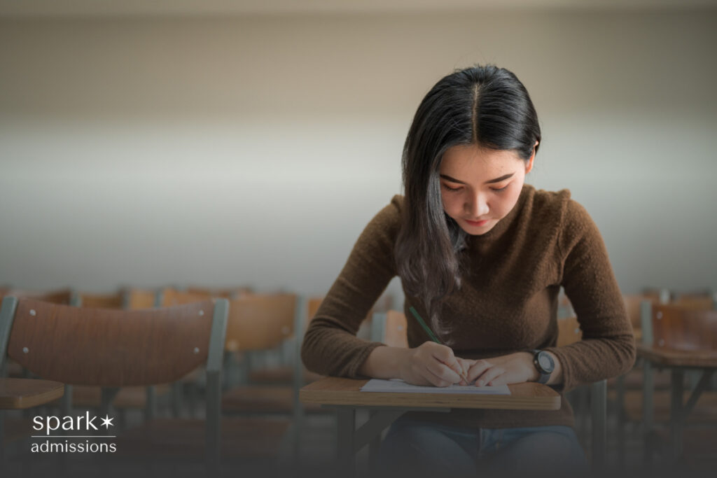 A young woman sits alone in a classroom filling out a paper on a wooden desk
