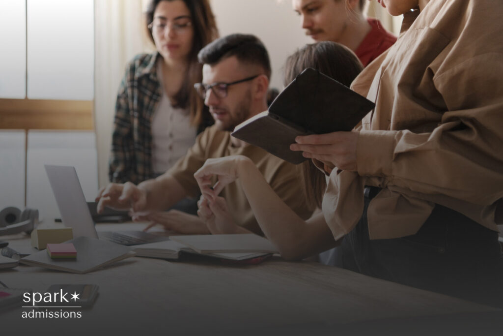 A group of young adults gathers around a laptop, discussing and pointing at the screen