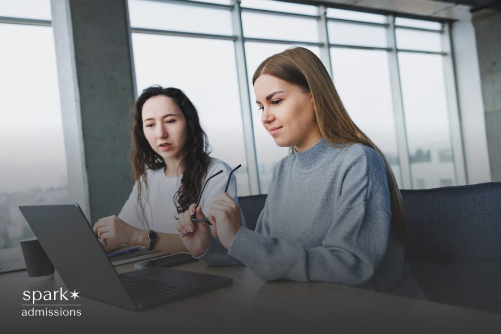 Two women engaged in a coaching session, with one guiding the other who is an intern, seated at a table with an open laptop in front of them displaying a work-related interface